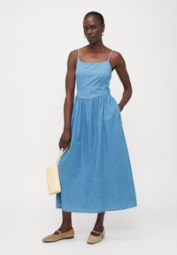 Vestido maxi de mezclilla azul con un torso ajustado y falda acampanada. Presenta tirantes delgados y una línea de cintura natural. Zapatos color tostado y un bolso amarillo.
