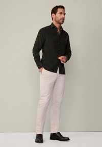 Camisa verde oscuro de botones, pantalones beige y zapatos negros. La camisa tiene cuello, diseño ajustado y textura suave. Fondo neutro.