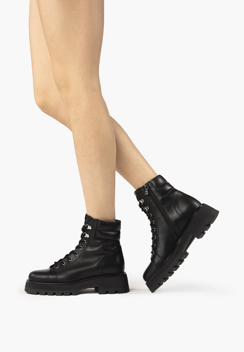 Black leather ankle boots with a lace-up front and silver hardware. Chunky sole with a textured pattern and a cuffed top design.