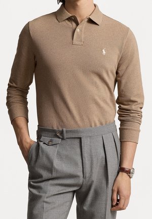 Homme portant un polo beige à manches longues rentré dans un pantalon gris taille haute à plis, avec une montre au poignet gauche.