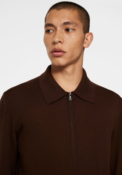Giacca a maglia marrone con colletto e zip frontale. Presenta una superficie texturizzata e un design aderente, che mette in risalto la parte superiore del corpo.