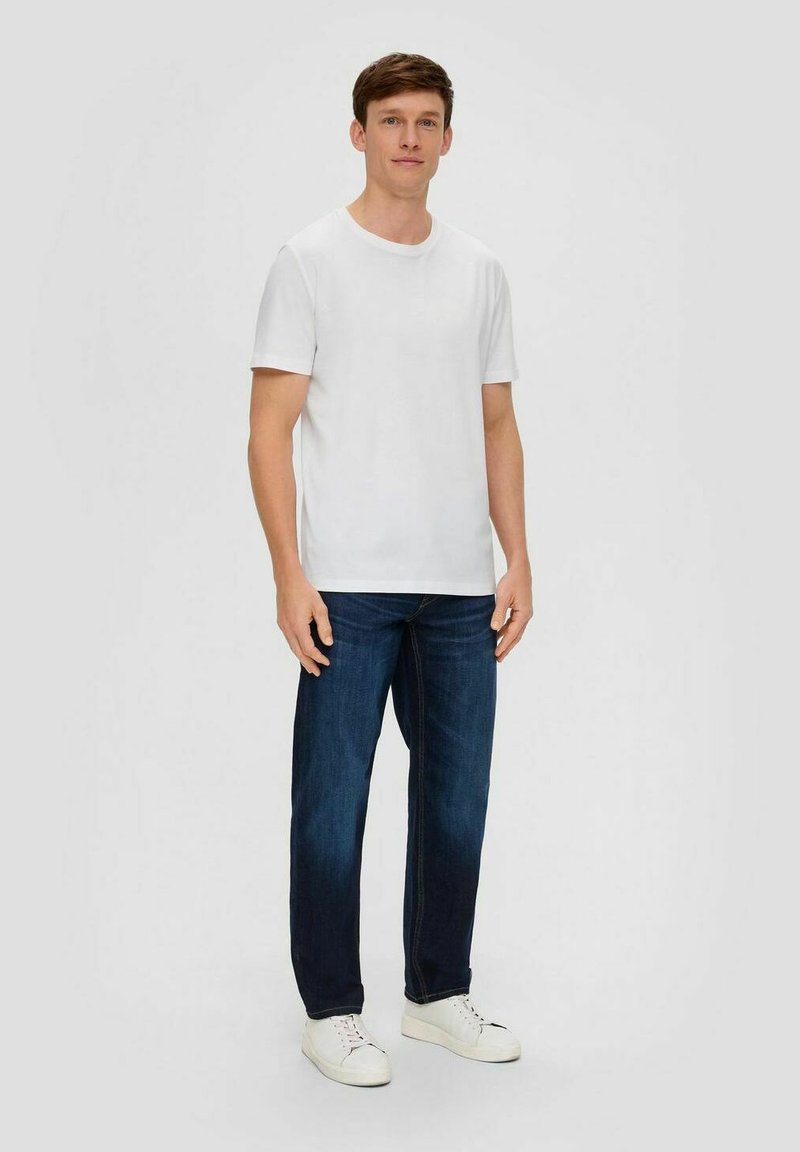 Weißes T-Shirt mit kurzen Ärmeln und Rundhalsausschnitt, kombiniert mit dunkelblauen Jeans und weißen Sneakers, stehend vor einem hellgrauen Hintergrund.