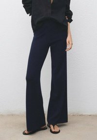 Persona che indossa pantaloni larghi blu navy, ciabatte nere e una maglietta nera a maniche lunghe, in piedi contro un muro bianco e testurizzato.