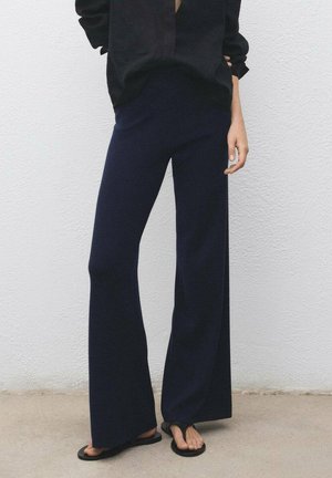 Persona che indossa pantaloni larghi blu navy, ciabatte nere e una maglietta nera a maniche lunghe, in piedi contro un muro bianco e testurizzato.