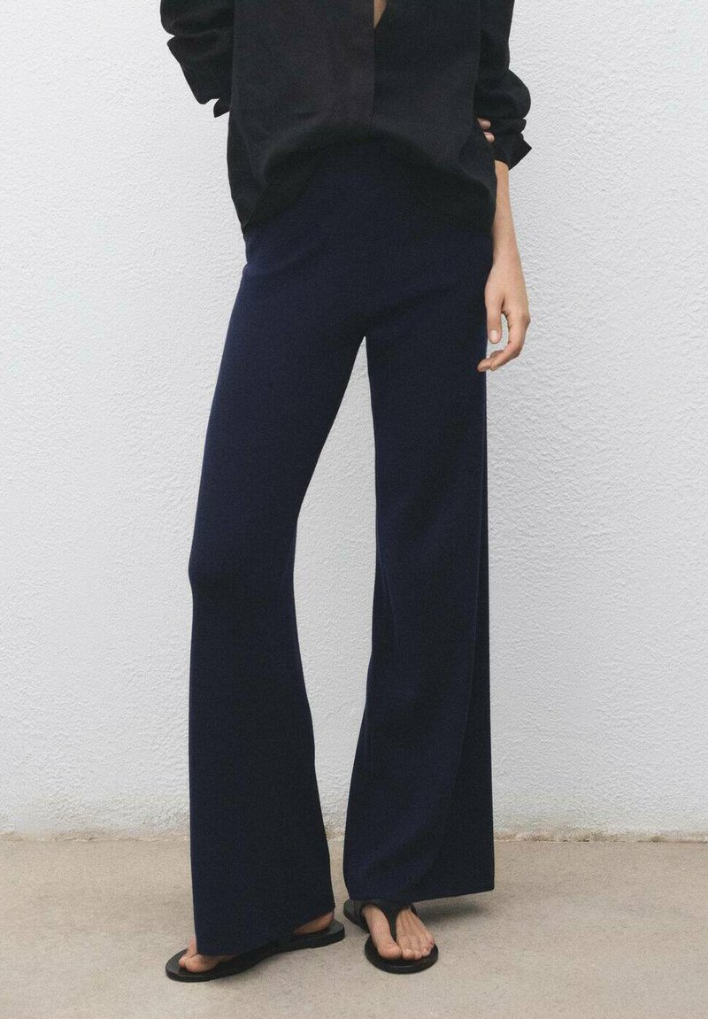 Persona che indossa pantaloni larghi blu navy, ciabatte nere e una maglietta nera a maniche lunghe, in piedi contro un muro bianco e testurizzato.