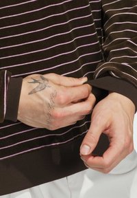 Brown striped sweater with pink lines. Close-up on hands adjusting sleeve cuff. Material appears soft, with a ribbed texture at hem and cuffs.