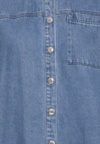 Tissu en denim bleu clair avec cinq boutons-pression métalliques au centre et une seule poche poitrine sur le côté droit.