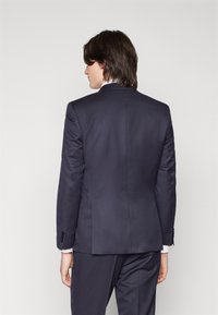 Marineblauwe colbert van gladde stof, in getailleerde pasvorm, met een enkele split, notch revers en accenten van vier knopen op de mouwen.
