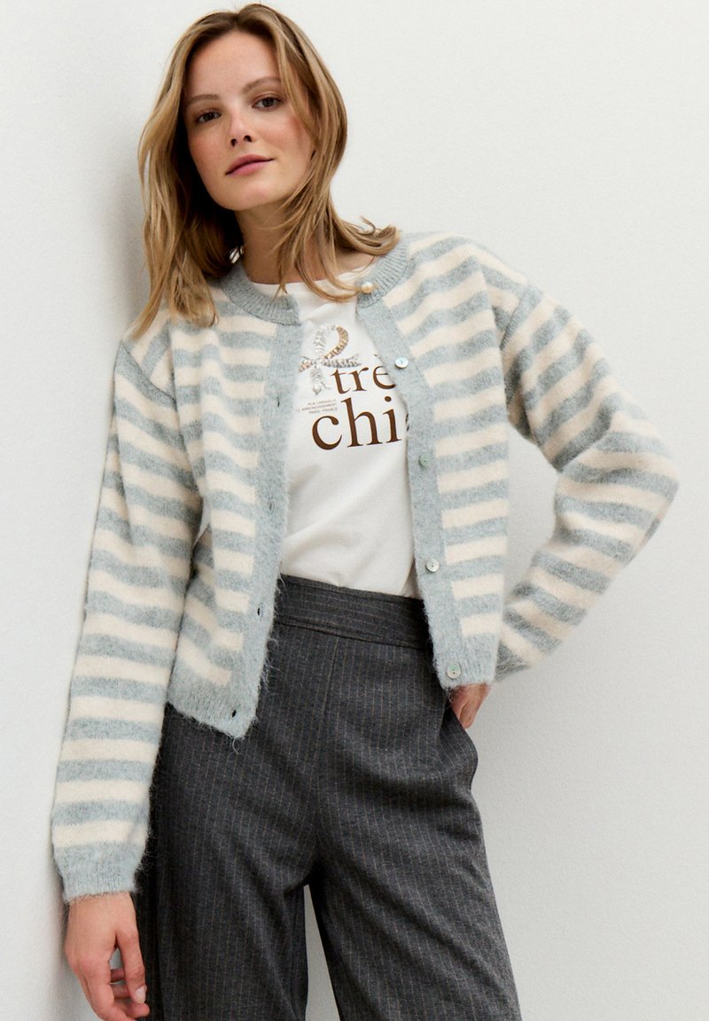 Eine Frau trägt eine hellgraue und weiße gestreifte, flauschige Strickjacke, ein graues T-Shirt mit Grafik und schwarze hochgeschnittene Hosen mit Nadelstreifen und steht gegen eine Wand.