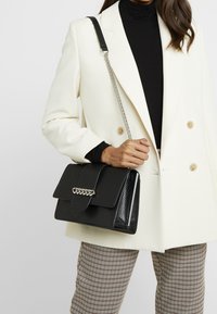 Borsa nera con un esterno texturizzato, dettaglio in catena argentata e chiusura a patta. Indossata con un blazer bianco e pantaloni a fantasia.