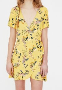 Robe portefeuille jaune avec manches courtes à volants, imprimé floral multicolore, décolleté en V profond et fermeture à nouer sur le côté gauche.