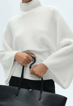 Person trägt einen weißen Rollkragenpullover mit weiten Ärmeln und hält eine schwarze Leder-Einkaufstasche an den Griffen auf Hüfthöhe.