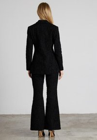 Zwarte gestructureerde kostuum met een getailleerde blazer en uitlopende broek. Het materiaal heeft een subtiel bloemmotief. Achteraanzicht toont de lengte en de vorm.