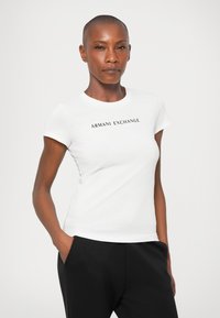 Bela kratka majica s kratkimi rokavi, narejena iz bombaža, s potiskom "ARMANI EXCHANGE" v črni barvi čez prsi. Gladka tekstura, priležen dizajn.