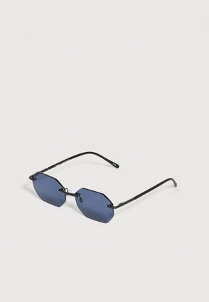Lunettes de soleil à monture noire avec des verres octogonaux bleu foncé et des branches fines reposant sur une surface claire.