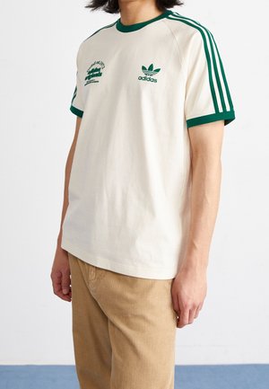 Osoba w białym T-shircie Adidas z zielonymi paskami na rękawach i zielonymi logo, w połączeniu z jasnobrązowymi sztruksowymi spodniami.