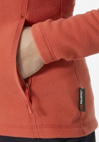 Veste en polaire rouge avec une poche zippée. La texture est douce et duveteuse. Comprend une étiquette Polartec sur le côté. Présente des poignets ajustés et un col haut.