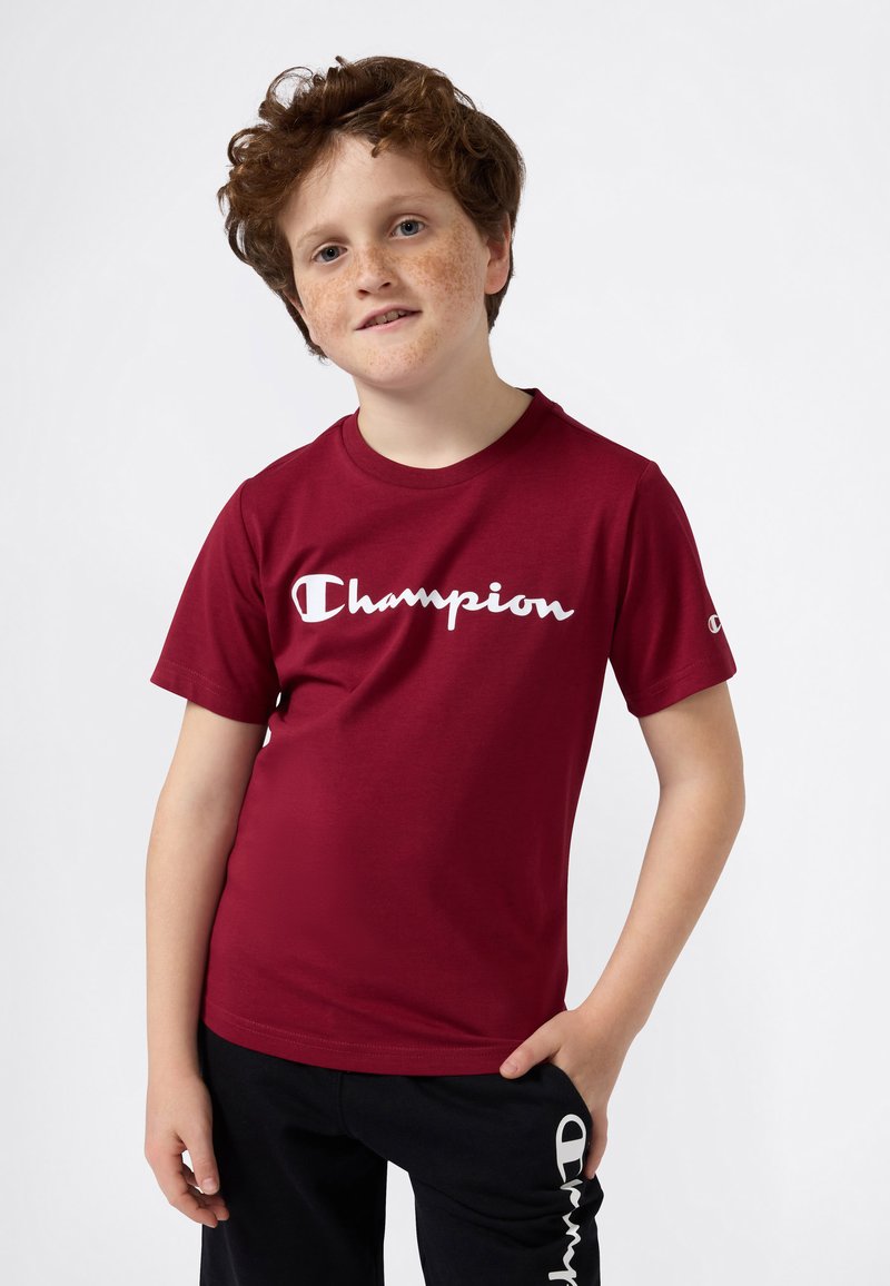 Röd bomullströja med korta ärmar, utrustad med en vit "Champion"-logga över bröstet och en rund halsringning. Matchas med svarta sweatpants.