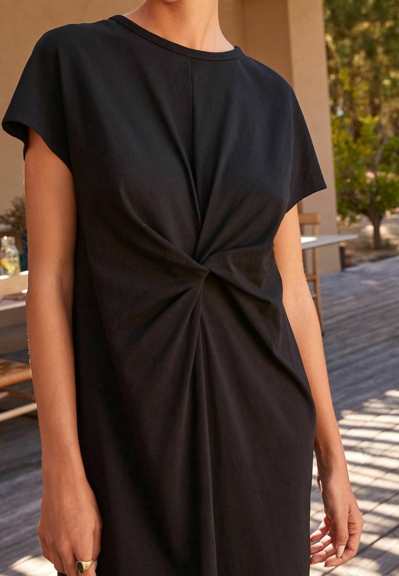 Femme portant une robe noire à manches courtes avec un détail de nœud tordu à la taille, se tenant à l'extérieur près d'une terrasse en bois et d'une table.