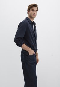 Hemd mit Naval-Kragen, aufgerollten Ärmeln, kombiniert mit dunklen Jeans. Das Hemd hat Knöpfe und wird über einem weißen T-Shirt getragen.