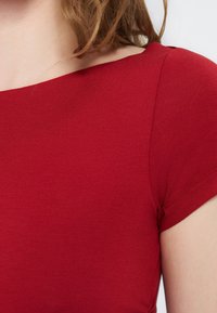 Κοντινό πλάνο ενός ατόμου που φορά εφαρμοστό κόκκινο κοντομάνικο τοπ με φαρδιά λαιμόκοψη και καστανά σγουρά μαλλιά.