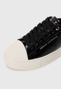 Baskets en cuir verni noir avec un embout en toile texturée blanche, lacets noirs et logo "Armani Exchange" en doré sur le côté et la semelle.