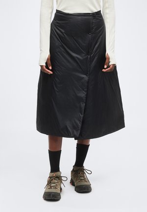 Personne portant une jupe midi matelassée noire avec fermeture par boutons, des chaussettes foncées, des chaussures de randonnée et un haut blanc à manches longues avec des trous pour les pouces.