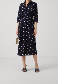 Donna che indossa un vestito midi a pois blu navy con una cintura, tiene in mano una borsa beige e indossa sandali con tacco blu decorati.