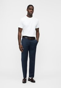 Mann steht und trägt ein weißes T-Shirt, marineblaue Hose, braunen Gürtel und braune Loafers vor einem schlichten hellen Hintergrund.