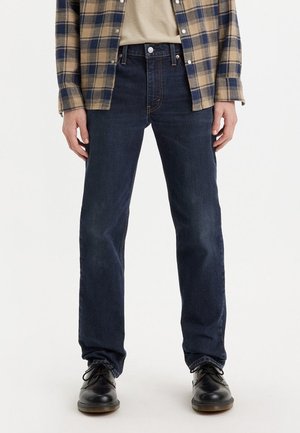 Jeans en denim bleu foncé avec une coupe droite, dotés de passants de ceinture et d'un design classique à cinq poches. Portés avec une chemise beige et une veste à carreaux.
