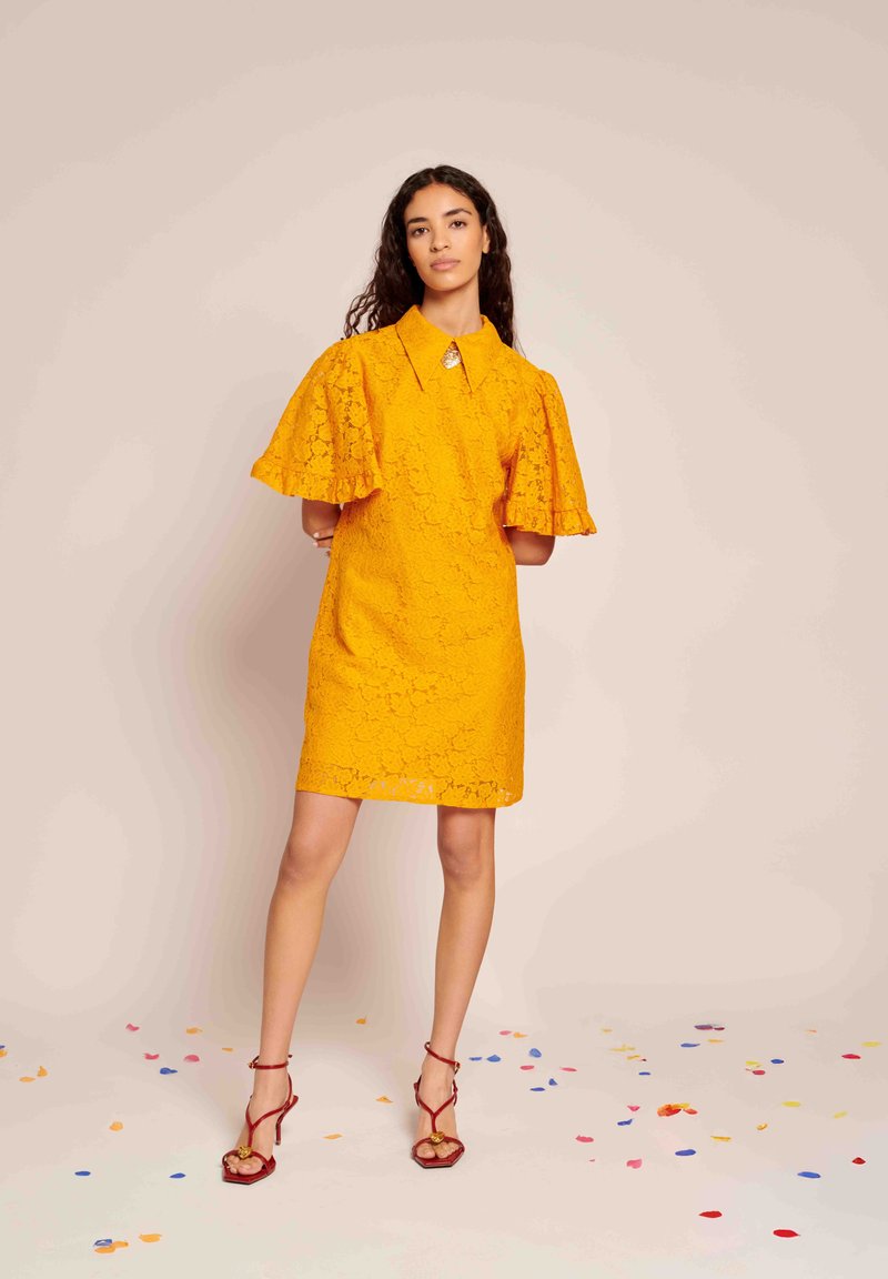 Vestido amarelo de renda com decote tipo colarinho e mangas largas. Apresenta um broche decorativo e é combinado com sandálias vermelhas de tira sobre um fundo de confete.