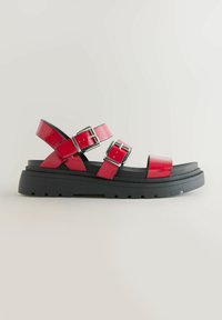 Sandales en cuir verni rouge avec deux sangles réglables et boucles en argent, dotées d'une semelle en caoutchouc noir épais et d'un profil plat.