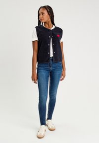 Gilet en polaire sans manches bleu marine avec un col rond, des fermetures à pressions et un design en forme de cœur rouge sur la poitrine, associé à un t-shirt blanc et un jean bleu.