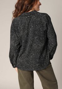 Zwarte blouse met wit abstract patroon, lange mouwen en een kraaghalslijn, gecombineerd met olijfgroene broek. Soepele stoftextuur.