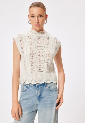 Top senza maniche lavorato a maglia color crema, con orlo a scallop, motivi a filigrana e design a collo alto. Abbinato a jeans in denim blu chiaro.