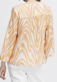 Donna che indossa una blusa lunga a maniche lunghe, larga, con motivi a spirale arancioni, rosa e bianchi, abbinata a pantaloni bianchi, vista di spalle.