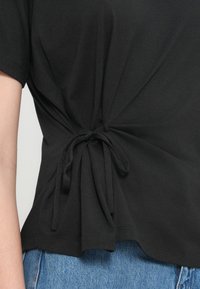 Μαύρο T-shirt από απαλό βαμβάκι, με λεπτομέρεια δέσιμου κόμπου στο πλάι, κοντά μανίκια και άνετη εφαρμογή. Φορεμένο με ανοιχτόχρωμο μπλε τζιν.