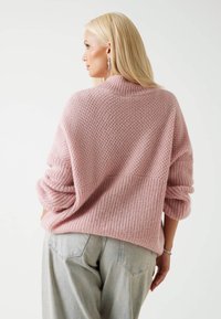 Ženska z dolgimi blond lasmi, oblečena v teksturirano roza pulover in svetle traper hlače, stoji s rokama v žepih in gleda s strani.