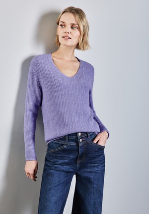 Lila gerippter Pullover mit V-Ausschnitt und langen Ärmeln, kombiniert mit dunkelblauen Jeans aus Denim mit einem geknöpften Bund und Fronttaschen.