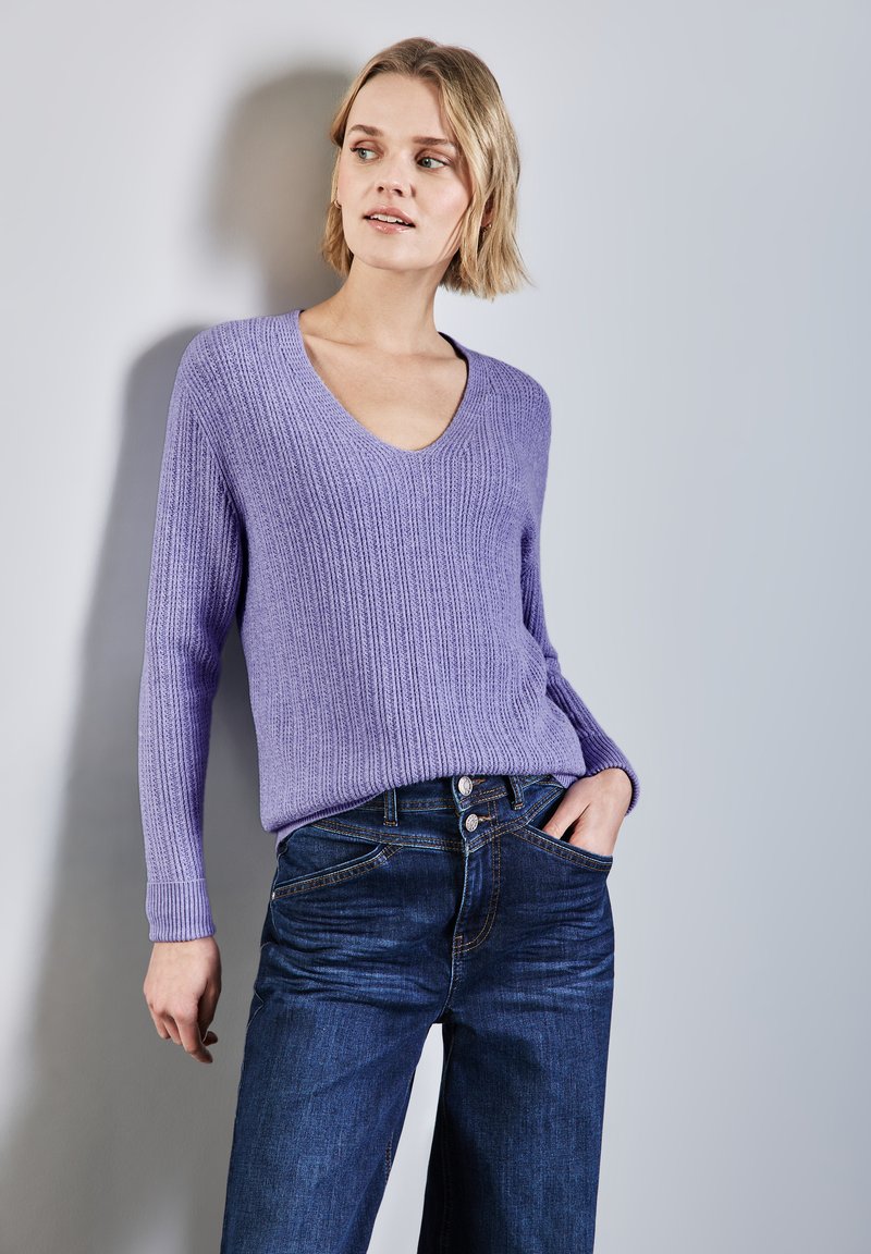 Lila gerippter Pullover mit V-Ausschnitt und langen Ärmeln, kombiniert mit dunkelblauen Jeans aus Denim mit einem geknöpften Bund und Fronttaschen.
