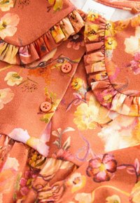 Tissu floral de couleur rouille avec des fleurs jaunes et roses, bords froncés, et deux petits boutons ronds orange sur le col d'un vêtement.