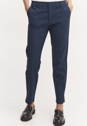 Marineblauwe broek met een slim fit, voorzakken en riemlussen, gemaakt van een gladde stof; gecombineerd met zwarte instappers.