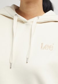 Sudadera crema hecha de tejido suave, con un bolsillo canguro, cordones con puntas de metal y el logo bordado de "Lee" en un color a juego.