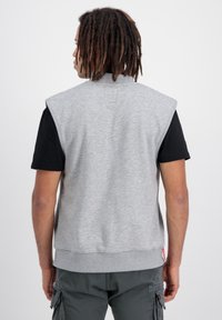 Grauer ärmelloser Hoodie aus weichem Material, mit geripptem Halsausschnitt, tiefen Armöffnungen und einem Saum mit kontrastierendem Nähen, getragen über einem schwarzen T-Shirt.