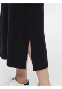Abito nero lavorato a maglia con spacco laterale, orlo a coste e texture morbida. Abbinato a una sneaker bianca con accentuazione nera sul tallone.