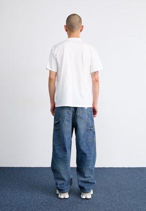 Person med barberet hoved, iført en plain hvid t-shirt og løse blå denim cargo bukser, stående med ryggen mod beskueren på blåt tæppe.