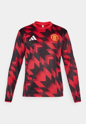 Langermet rød genser med svarte geometriske mønstre, med et brodert Manchester United-logo og tre hvite striper på skulderen.