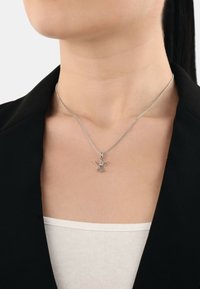 Collana con pendente d'argento che presenta un dettagliato design di angelo su una catena sottile, indossata sopra una maglietta bianca e sotto un blazer nero.