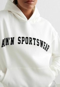 Sudadera blanca con un destacado gráfico curvado en negro que dice "AIM'N SPORTSWEAR". Cuenta con capucha y bolsillo frontal, textura de tela suave.