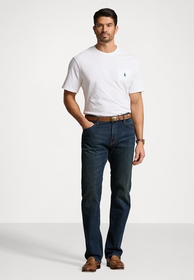White cotton t-shirt with pocket, dark blue denim jeans, brown leather belt, and loafers. Model stands with hands in pockets against a plain background.